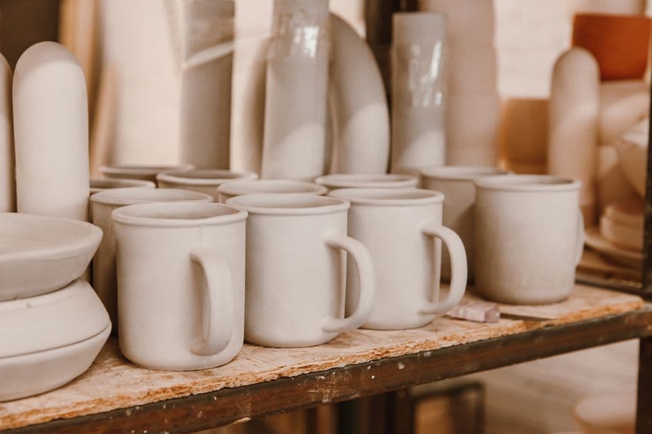 Rows of handmade cream stoneware mugs drying on a studio shelf in warm light